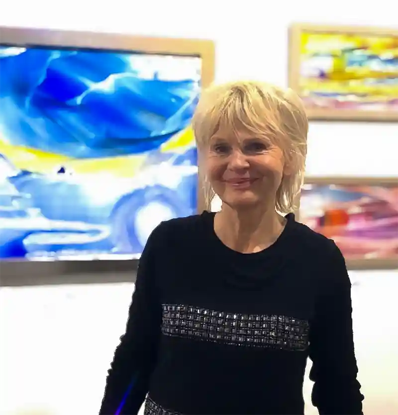 Portrait Sylvia Sanders, lächelnd in die Kamera schauen, blonde Haaren steht vor eigenen Kunstwerken