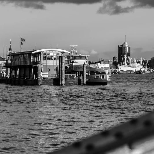 Schwarz weiß Foto über die Elbe, im Hintergrund die Elbphilharmonie und HTC, im Vordergrund ein unscharfer Brückenpfeiler, Detaiansicht mit Fähre