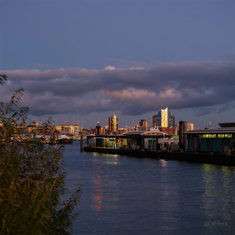 Farbfotografie des Hafens, das Abendlicht spiegelt sich in der Fassade der Elbphilharmonie, leichte Wolken am Himmel. Detailansicht mit Elbphilharmonie