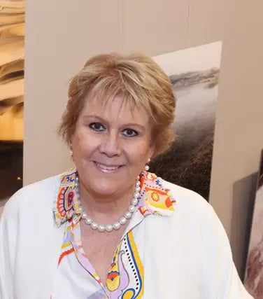 Die Künstlerin schaut frontal in die Kamara, helle Jacke, lächelt vor wand mit eigenen Kunstwerken